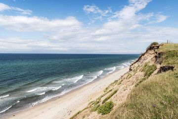 A scenic coastal landscape featuring a sandy beach bordered by gentle waves and a deep blue sea. A grassy, sunlit cliff along the shoreline under a partly cloudy sky, peaceful and natural seaside
