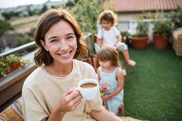 Close-up of smiling parent holding coffee cup, beige bench and green lawn in soft focus, terrace with plants behind, warm light, mood of calm joy and family connection.