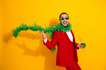 Funny stylish man in red suit with green feather boa smiles and party ready holding a green drink against bright yellow background for bold fashion and humor vibes