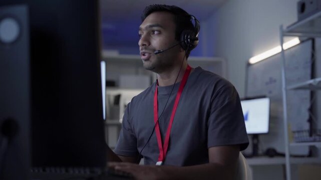Remote work online technical support concept. Portrait young adult indian man wearing headset typing keyboard working in office. It support business customer service technology.