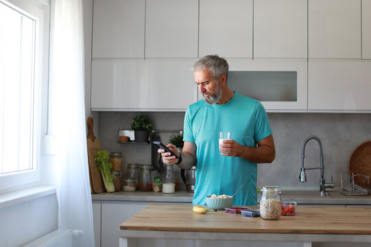 Portrait of a happy senior mature mid aged man having a healthy breakfast after fitness exercise training and using a smartphone, looking at phone and holding a glass of milk in the morning at home - Powered by Adobe