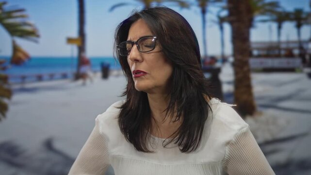 Middle aged hispanic woman clutch chest on street by seaside promenade lined with palm trees under sunny sky; heartattack pain.