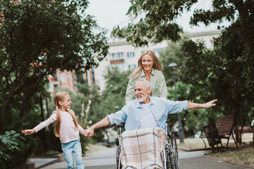 Fototapeta premium Joyful family moment as grandpa in wheelchair is cared for by daughter and wife in a sunny park walking together near trees and benches