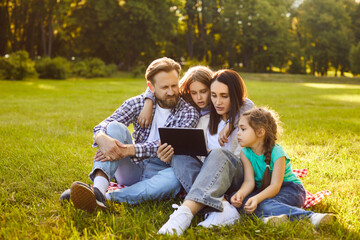 Fototapeta premium Happy family using tablet, sitting together on green grass of summer park. Mom and dad, kids hugging while reading digital book or text on modern device screen. Family picnic, bonding concept
