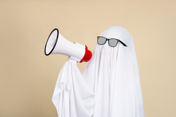 Funny Halloween ghost wearing white cloth shouting into megaphone isolated on beige background. Happy Halloween or weird concept.