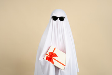 Halloween ghost in white cloth holding gift box expressing happy isolated on beige background. Happy Halloween concept. Crazy idea.