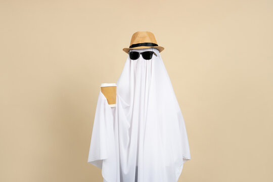 Person in a white ghost costume wearing sunglasses and a straw hat holding a cup of hot coffee on a beige background.