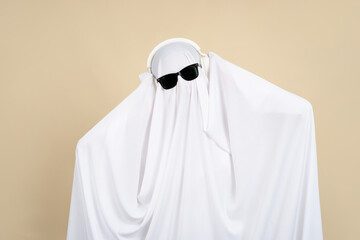 Cool ghost, man wearing sunglasses and headphones covered with a white cloak on a beige background.