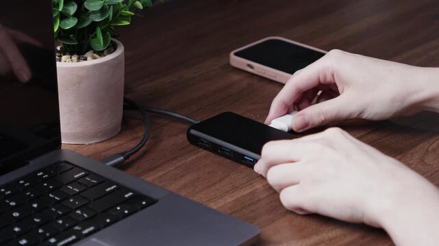 Person connecting a white USB-C cable to a black multi-port hub, expanding laptop connectivity on a modern wooden desk for a productive digital workspace
