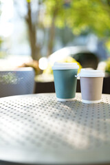 Two coffee cups sit on a metal table in a sunny outdoor cafe setting