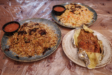 A high-angle view of a delicious Middle Eastern or Arabic feast. Two large plates of aromatic Mandi or Biryani rice, topped with crispy fried onions, are served alongside a dish of roasted meat. 
