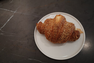 A close-up, high-angle view of a golden-brown, flaky croissant generously topped with toasted sesame seeds. The pastry is served on a simple white ceramic plate, which sits on a dark, elegant, veined 