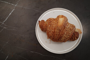 A close-up, high-angle view of a golden-brown, flaky croissant generously topped with toasted sesame seeds. The pastry is served on a simple white ceramic plate, which sits on a dark, elegant, veined 
