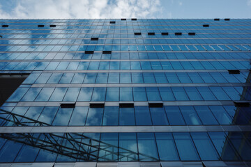 A low-angle, abstract view of a modern blue glass facade reflecting a cloudy sky.
