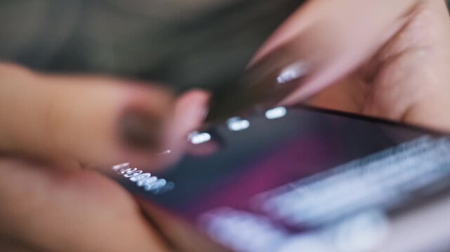 Close-up side view of a woman's hands with a brown manicure scrolling quickly through a social media feed on a smartphone. Her thumb rapidly swipes across the illuminated touch screen.