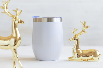 Blank Stainless Steel wine tumbler near golden Christmas deers on white table closeup, mockup