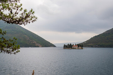 The small island of Saint George with its Benedictine monastery stands quietly in the Bay of Kotor, surrounded by green hills under cloudy skies.