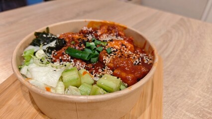 Korean rice bowl topped with spicy sauce, vegetables, sesame seeds, and green onions served in a paper container. The image represents Asian street food, bold flavors, and modern casual dining