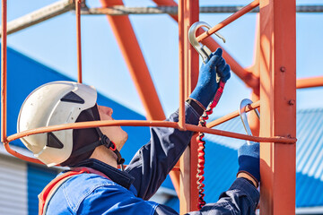 A construction worker in safety gear is installing equipment on a tall structure. The setting is bright and industrial, emphasizing safety and diligence.