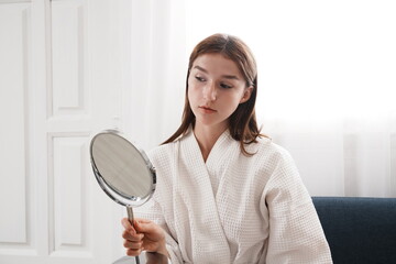  young woman in a bathrobe sits holding a mirror, looking at her fresh, clean, and nourished skin before applying makeup in her bedroom at home.