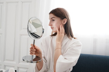  young woman in a bathrobe sits holding a mirror, looking at her fresh, clean, and nourished skin before applying makeup in her bedroom at home.