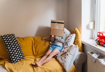 Emotional escape. Little child sitting on sofa at home with a cardboard box on head hiding from outside world and family conflict. Screen-free, safety childhood concept. Escape from overconsumption.
