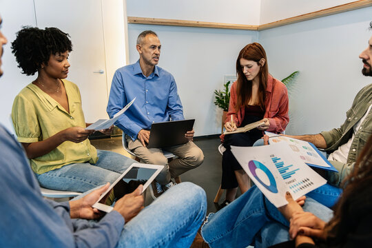 Group of coworkers discussing charts and ideas during a relaxed office meeting. Multicultural professionals collaborating in a creative environment. Teamwork concept. - Powered by Adobe