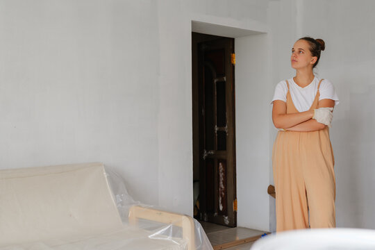 A woman stands thoughtfully in a nearly empty room with white walls. She wears overalls and appears to consider her home renovation options