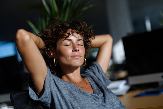 A woman with curly hair gently leans back in an office chair, basking in sunlight with eyes closed, capturing the essence of relaxation and personal well-being in a modern workspace.