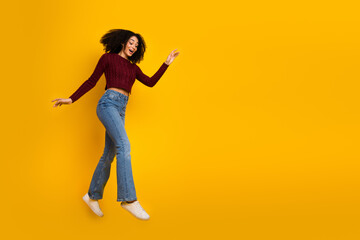 Joyful young mixed race woman jumping in a burgundy sweater over a bright yellow background promoting casual fashion and upbeat lifestyle