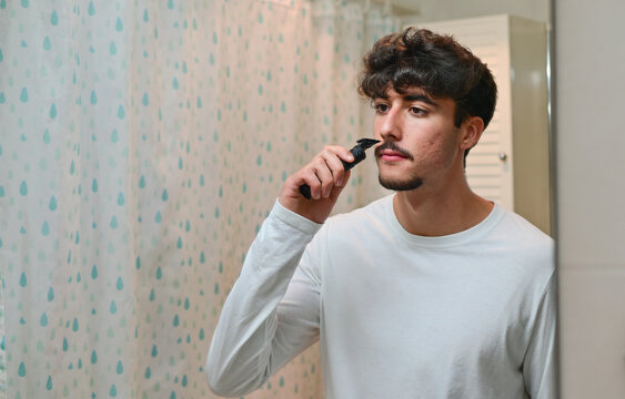 attractive young man shaving beard with electric shaver while looking at mirror in bathroom