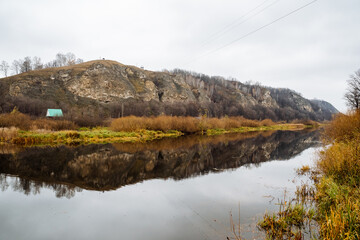 riverside cottage under sandstone ridge, quiet water reflects steep textured bank and sparse trees in subdued autumn light, remote retreat mood for slow travel and escape