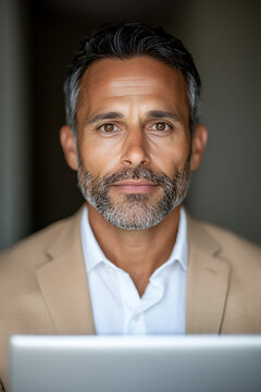 Portrait of a mature man with a stylish beard, wearing a jacket and a shirt. The background is dark. He is looking at the camera with a gentle expression.