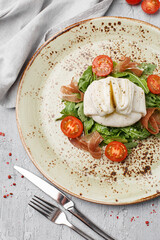 Salad with burrata cheese, arugula, cherry tomatoes and prosciutto on gray concrete background. Semisoft white italian cheese made from mozzarella and cream. Overhead view