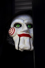 Frightening puppet mask with glowing green eyes and red spiral cheeks symbolizing horror and mystery in dark cinematic atmosphere.