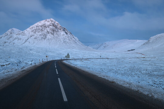 A night winter landscape in the Scottish Highlands, Glencoe. The snow-dusted A82 road curves towards the iconic, snow-capped mountain of Buachaille Etive Mor under a cold blue sky.  - Powered by Adobe