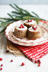 Three mini Christmas puddings decorated with cranberries and sugar powder on white festive plate with fir branches and red napkins over white wooden background. Close up view