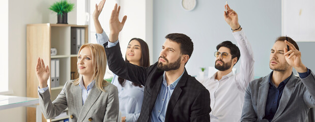 Young colleagues and company employees raising hands to vote at the conference sitting in a row in meeting room. Confident business people men and women voting in office during a meeting. Banner.