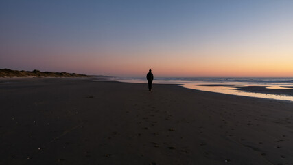 Naklejka premium Silhouette of a person standing alone on a beach at sunset. Lone figure watching the ocean during twilight. Solitude and contemplation concept.