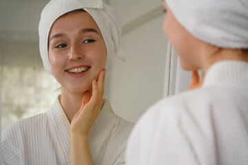 beautiful young woman wearing a bathrobe and using a towel to cover her hair after taking a shower looks at her face in the mirror and applies facial cream in her dressing room at home