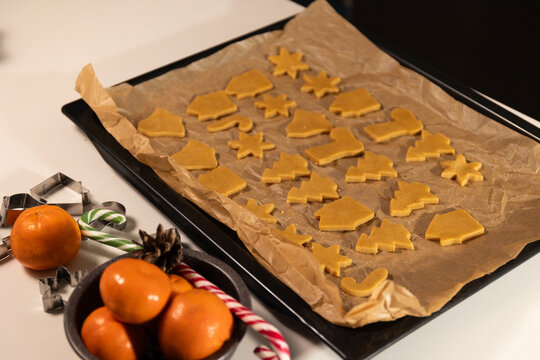 Gingerbread cookies in Christmas shapes are being prepared on a baking sheet. A bowl of tangerines and candy canes are visible. Baking during the Christmas season is a traditional activity.