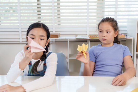 Asian preteen girl holding pink origami airplane in hand while caucasian classmate looks on, both engaged in classroom craft activity learning creativity, teamwork, and fun childhood skill - Powered by Adobe