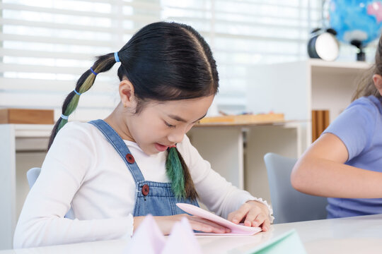 Asian preteen girl in denim overall carefully folding paper sheet into origami shape during classroom craft activity, practicing creativity and patience as part of afterschool learning project - Powered by Adobe