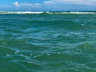 A powerful shot of the deep green-blue ocean with a series of rolling waves and white foam on the surface under a vast blue sky.