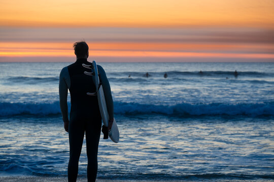 As the sun sets over Courant de Huchet, a surfer stands by the shore, gazing at the waves, with fellow surfers riding the swells in the distance. The sky glows with vibrant colors. - Powered by Adobe