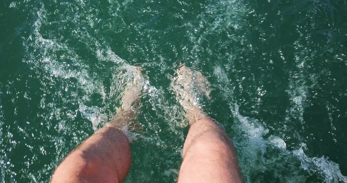 Caucasian male's legs and submerged feet in clear, turquoise water, creating splashes and ripples in the wake of a small rental powerboat stern deck. POV perspective, looking down, real time footage 