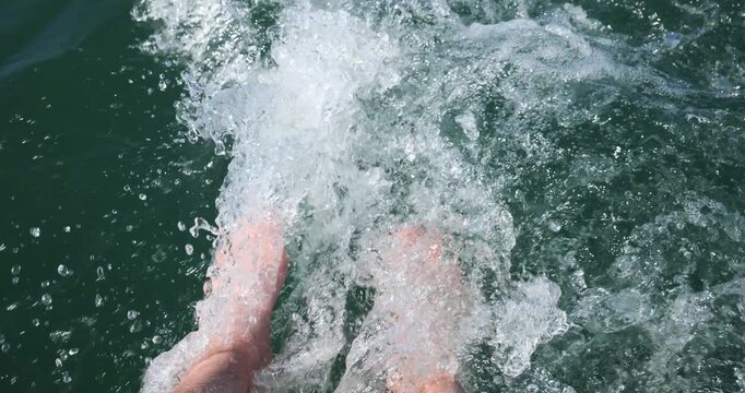 Caucasian male's legs and submerged feet in clear, turquoise water, creating splashes and ripples in the wake of a small rental powerboat stern deck. POV perspective, looking down, real time footage 
