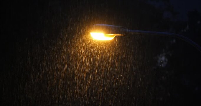 Close-up shot of a street lamp illuminating falling raindrops during heavy rain at night. Bright orange light, dark, moody atmosphere, real time footage, no people, elevated point of view