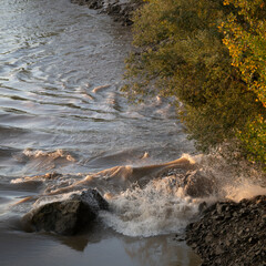 Waves crash onto rocky shores as water flows during the Vague mascaret at sunset in Podensac, Gironde. The scene captures the beauty of nature as the waves rise and fall.