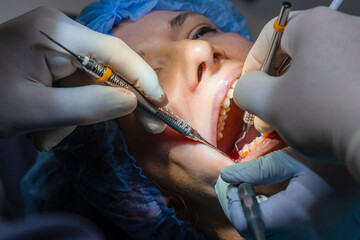Dentist performs procedure in woman mouth using instruments to gain access and manipulate area. Assistant maintains ensuring vision during treatment. 
